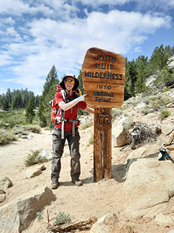 Linda Curtis in backpacking gear, smiling and pointing to a sign that says John Muir Wilderness.