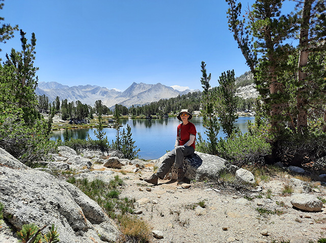 Linda Curtis sitting on a boulder in front of a body of still water, with mountains visible in the distance.