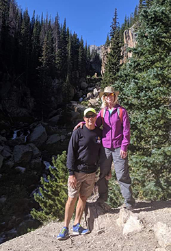 Linda Curtis and husband in front of a landscape with lots of pine trees and boulders.